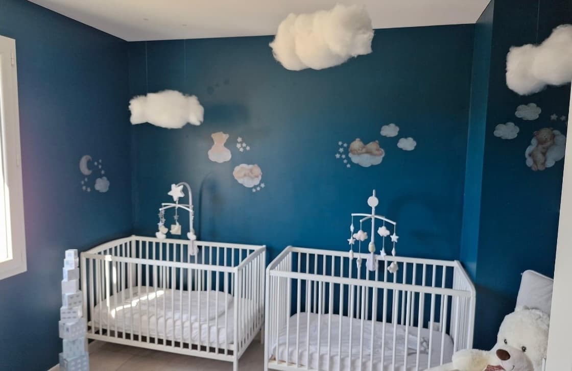 Une chambre de sieste chaleureuse avec deux lits à barreaux blancs sous un ciel bleu décoré de nuages suspendus.