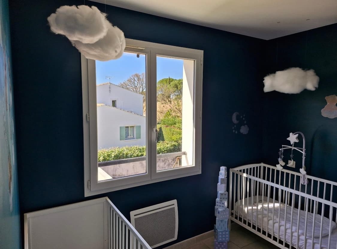 Une chambre de sieste chaleureuse dans une MAM, avec des lits à barreaux blancs, des murs bleu nuit et des nuages suspendus au plafond.