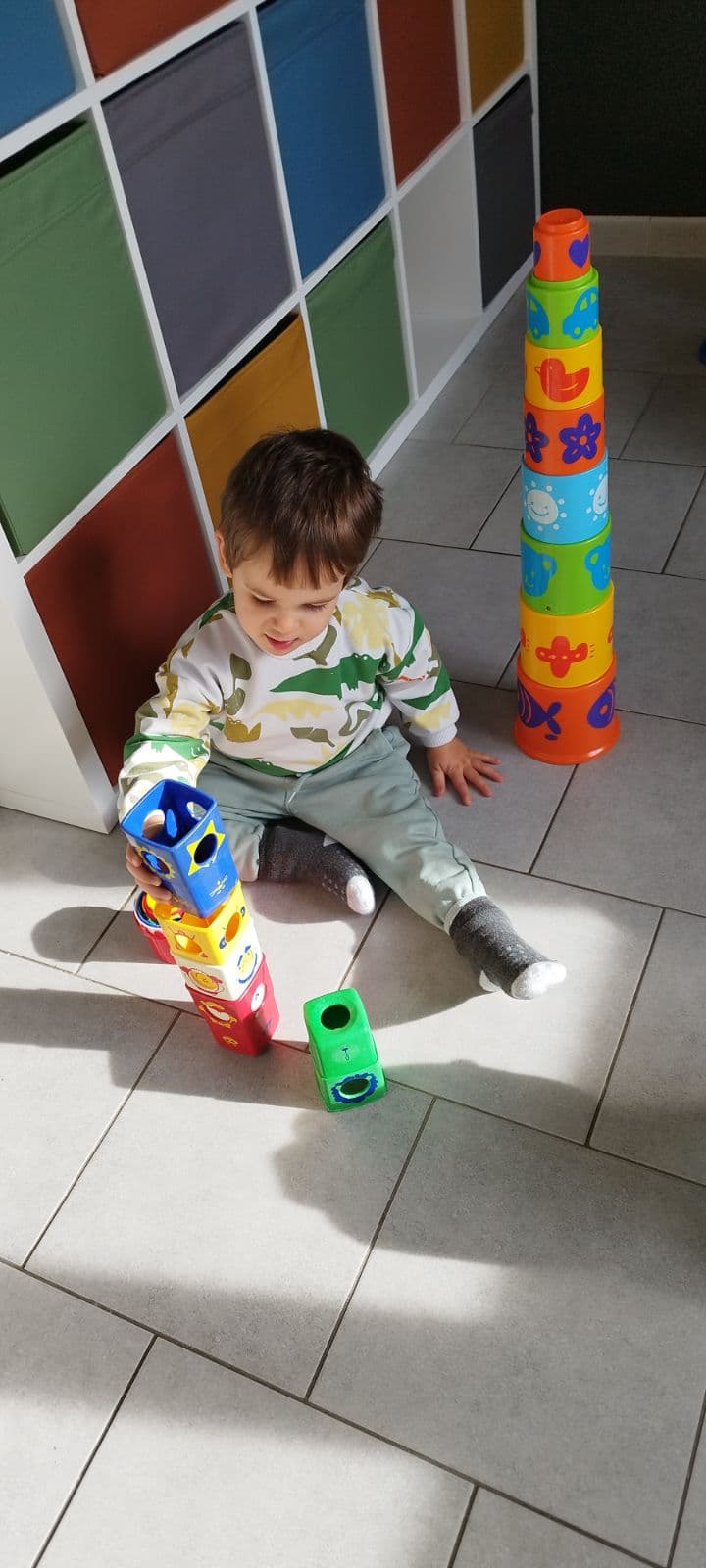 Un enfant assis au sol joue à empiler des cubes colorés à côté d'une grande tour de gobelets multicolores.
