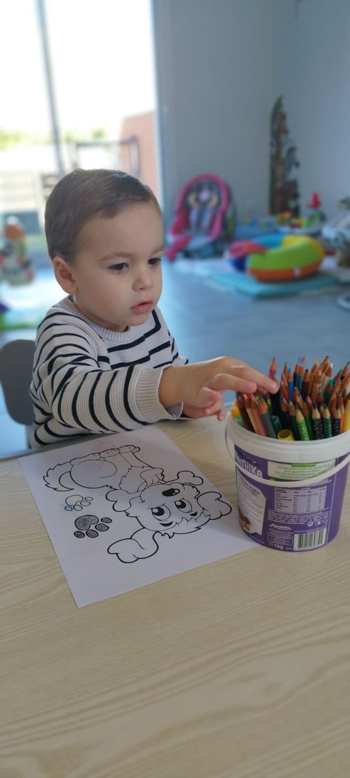 Un enfant assis à une table en bois choisit un crayon de couleur dans un pot pour colorier un dessin de chien.
