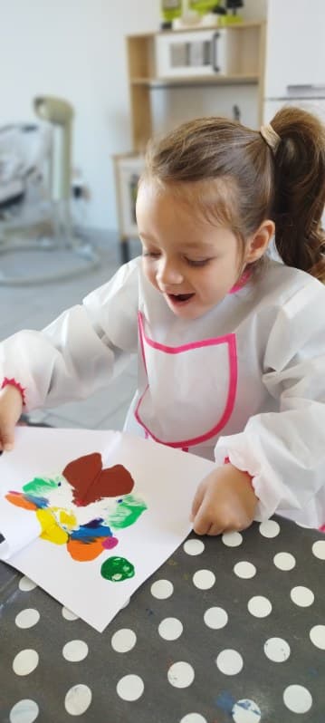 Un enfant portant un tablier de protection regarde avec enthousiasme sa peinture colorée sur une feuille de papier posée sur une table.