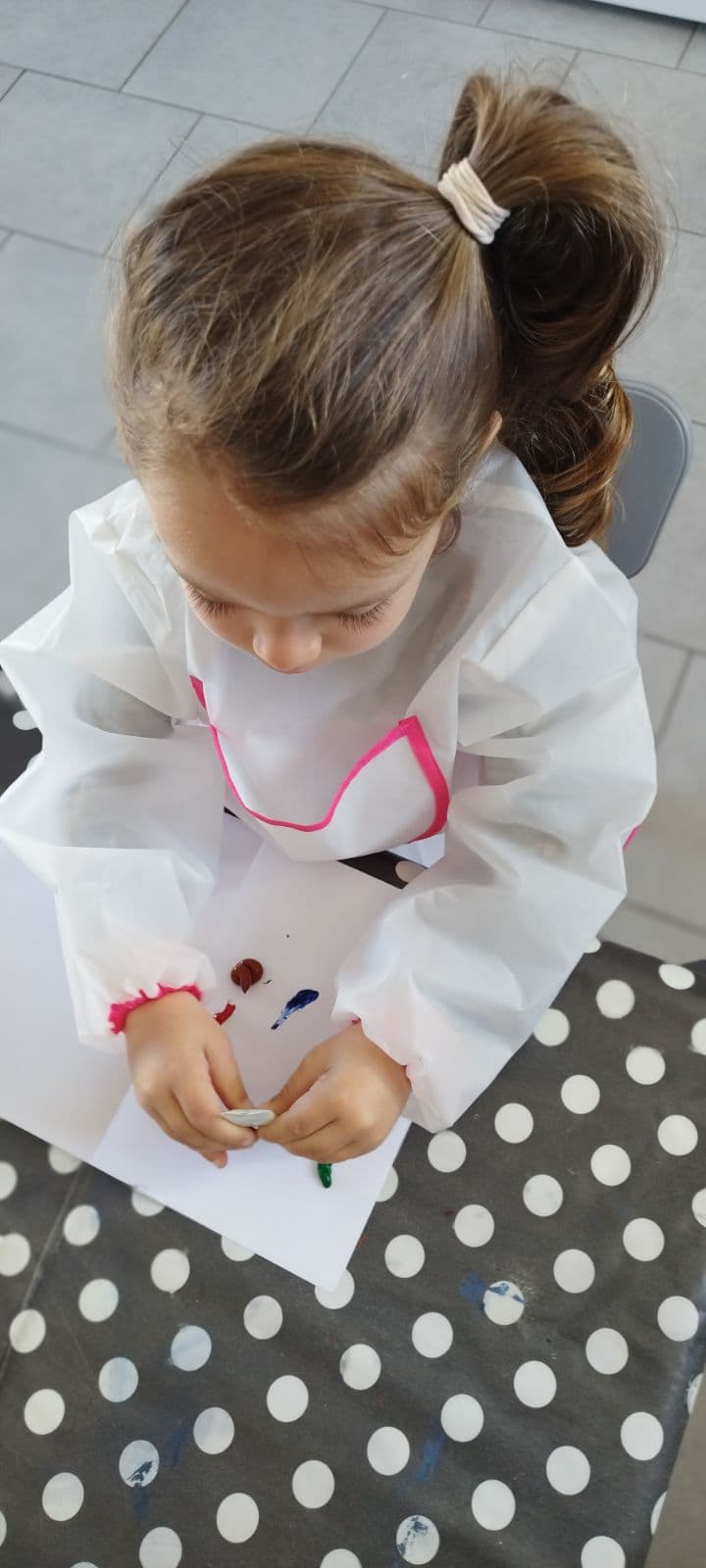 Un enfant portant une blouse de protection est concentré sur une activité manuelle, manipulant de la pâte à modeler sur une feuille de papier.
