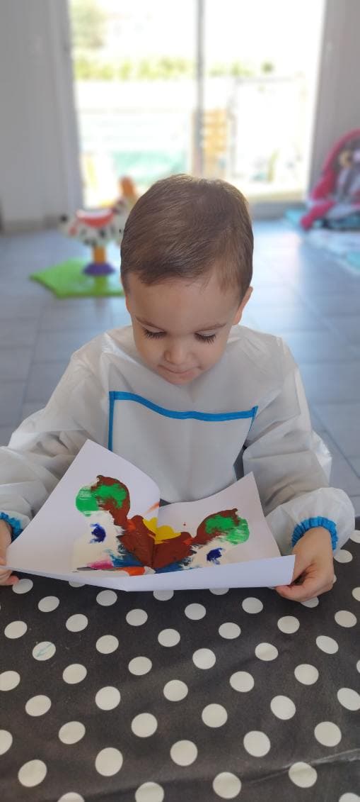 Un enfant assis à une table présente sa peinture colorée réalisée sur une feuille pliée en deux.