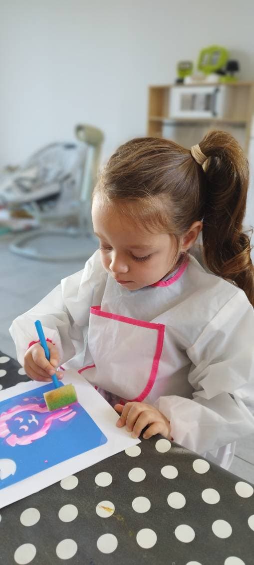 Un enfant concentré utilise un pinceau éponge pour peindre sur une feuille de papier posée sur une table à pois.