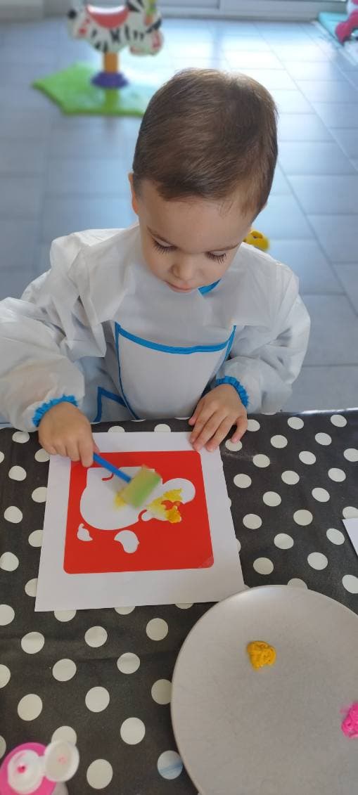 Un enfant portant un tablier de protection utilise un pinceau éponge pour appliquer de la peinture jaune sur un pochoir rouge posé sur une table.