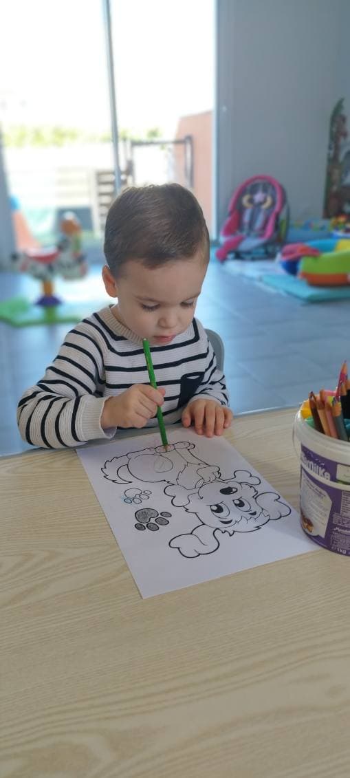 Un enfant concentré assis à une table en bois colorie soigneusement un dessin de chien avec un crayon vert.