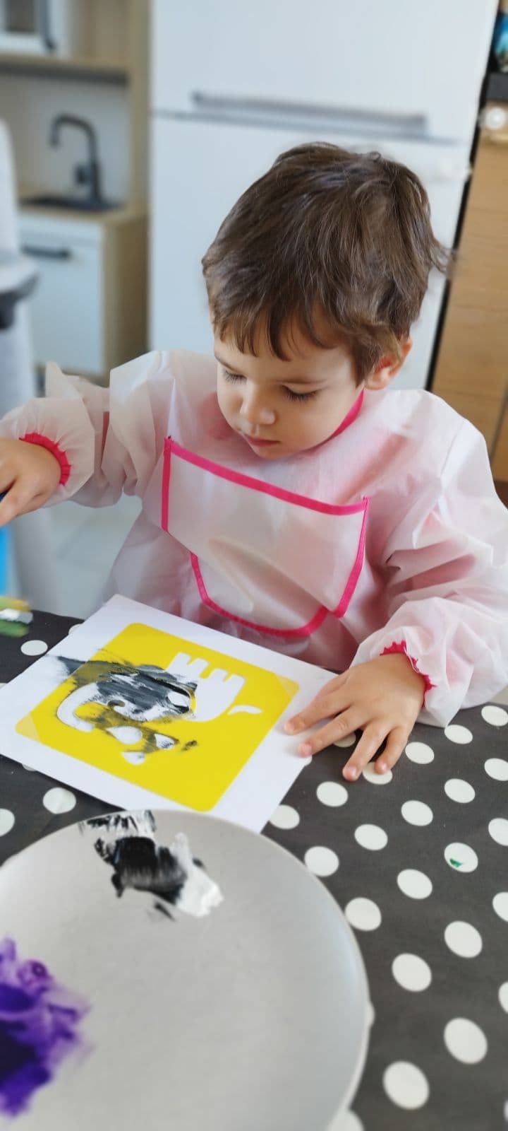 Un enfant porte une blouse de protection et réalise une activité de peinture avec un pochoir sur une feuille blanche, concentré sur son œuvre artistique.