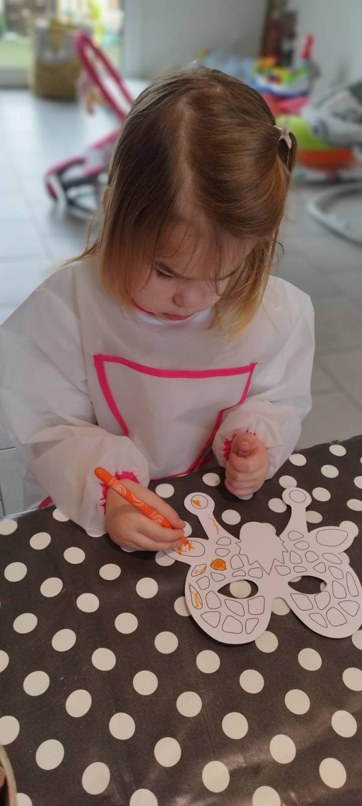 Un enfant concentré colorie un masque en papier en forme de girafe avec un feutre orange, portant une blouse de protection pour ses activités créatives.