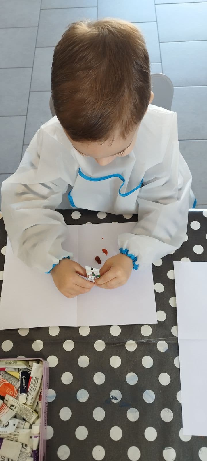 Un enfant portant une blouse de protection manipule un tube de peinture pour réaliser une activité artistique sur une feuille blanche posée sur une table.