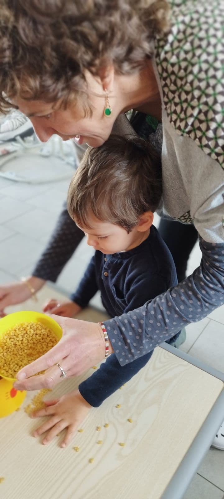 Une assistante maternelle accompagne un enfant dans une activité de manipulation avec des petites pâtes sèches sur une table.