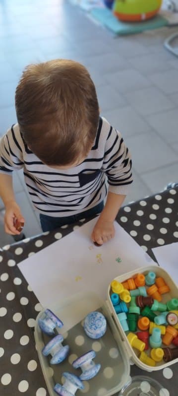 Un enfant utilise des tampons encreurs colorés pour créer des motifs sur une feuille de papier blanche, assis à une table avec des outils de création.