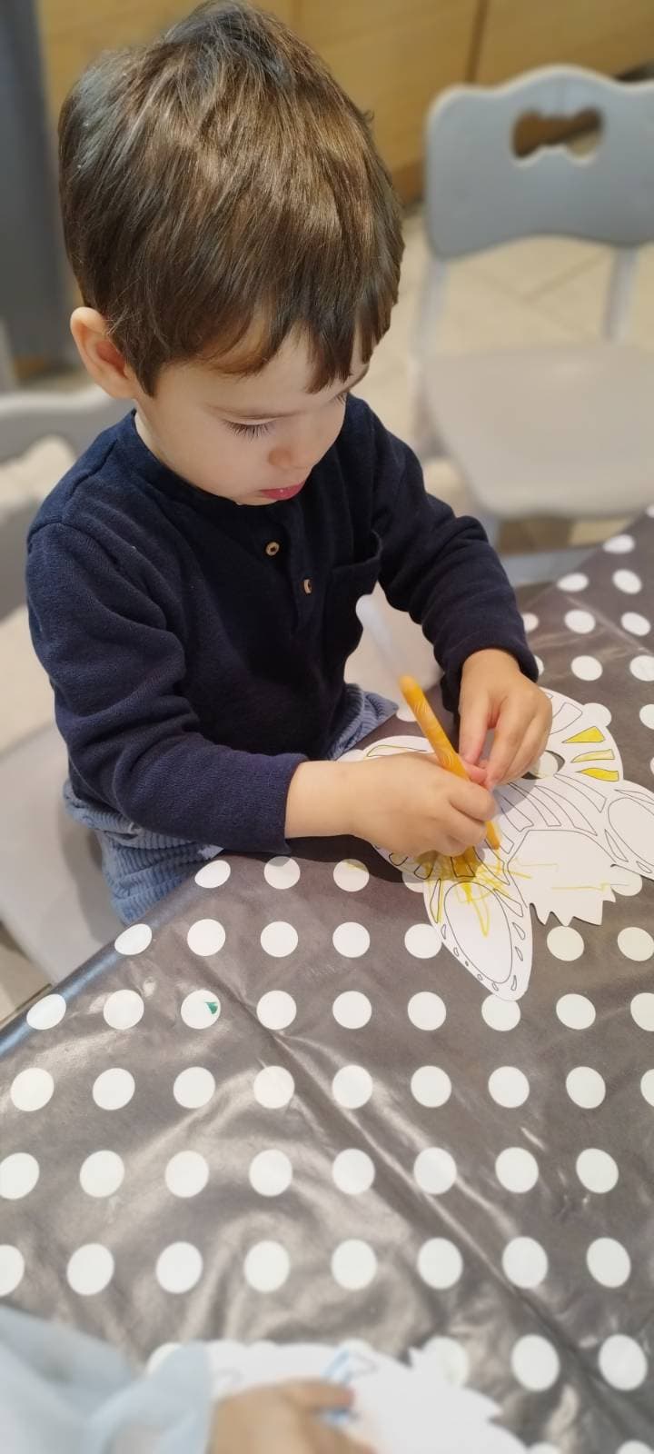 Un enfant concentré utilise un feutre jaune pour colorier un dessin sur une table recouverte d'une nappe à pois.