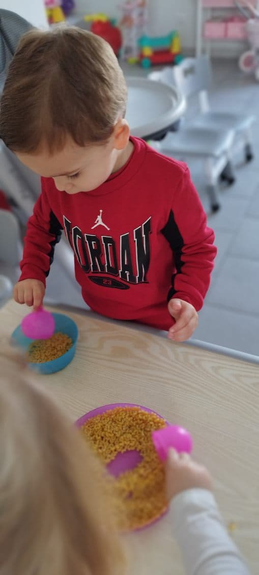 Des enfants jouent avec des petits récipients et des céréales sur une table, explorant les textures et développant leur motricité fine dans une ambiance ludique