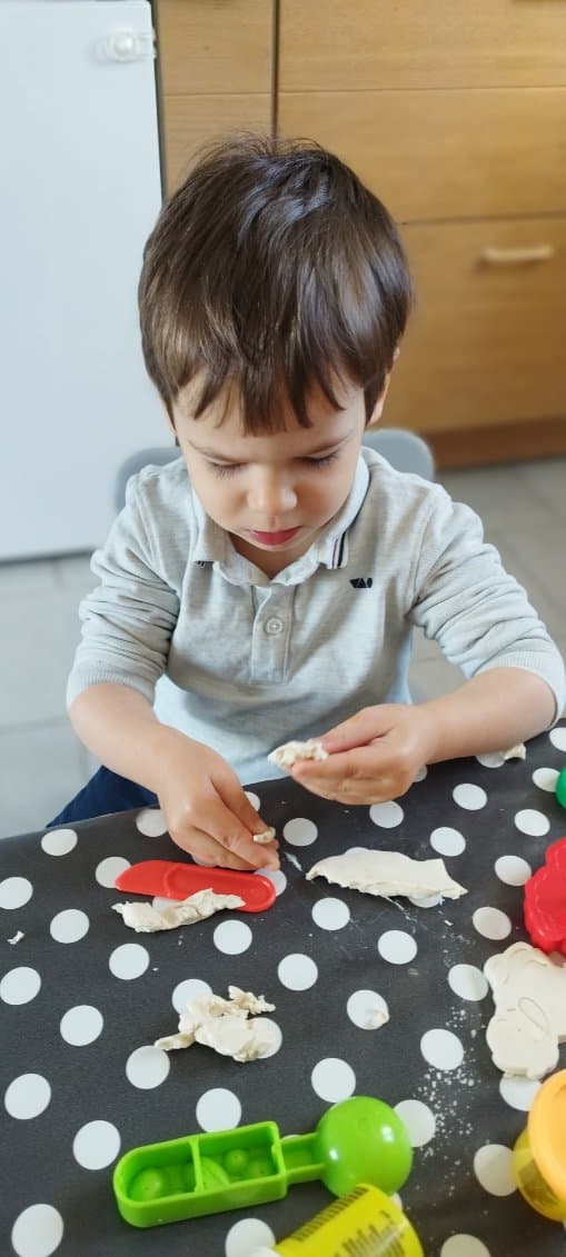 Un enfant concentré manipule de la pâte à modeler sur une table recouverte d'une nappe à pois, entouré d'outils de modelage colorés.