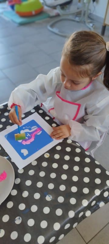 Un enfant concentré réalise une activité de peinture avec un pinceau mousse sur une feuille posée sur une table à pois.