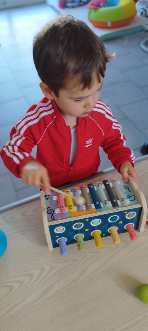 Un enfant en veste rouge joue avec un xylophone en bois coloré, explorant les sons et les notes avec une petite baguette.